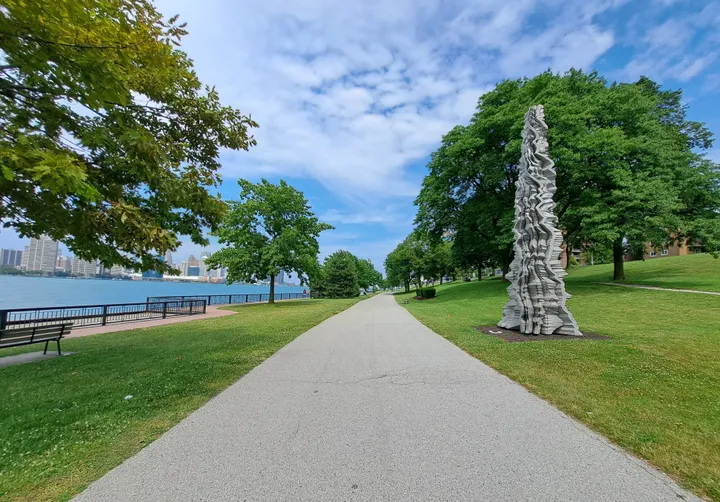 Central paved pathway surrounded by trees and sculpture along the Detroit river